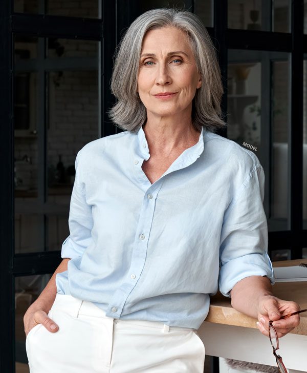 Middled aged female model leaning on desk with one hand in her pocket and the other hold glasses